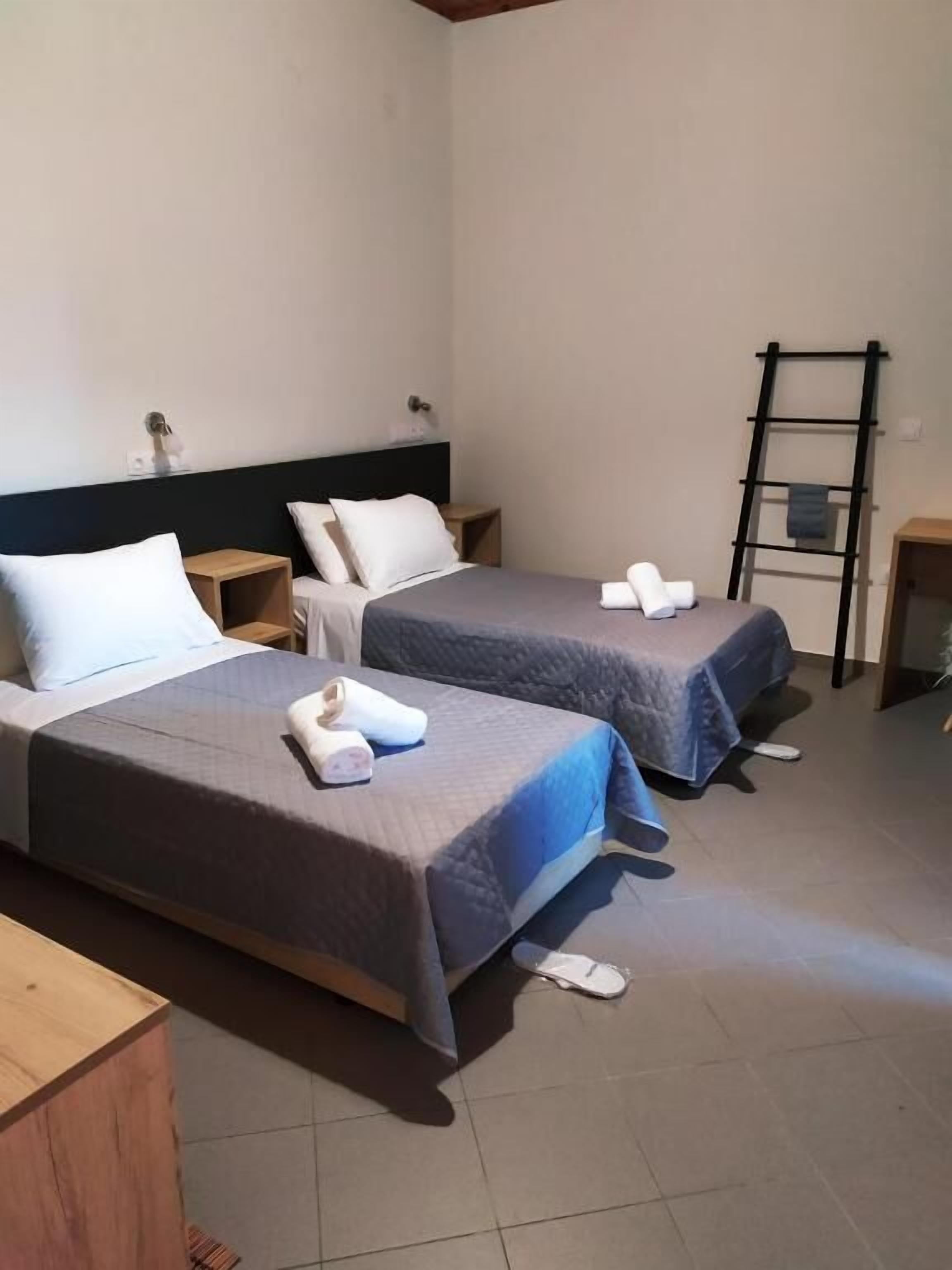 A hotel room with two beds, each with a pillow and a rolled towel. There is a wooden nightstand with a lamp, a ladder against the wall, and a small table with a towel.