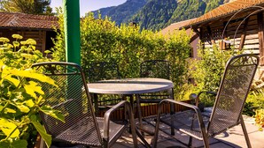 Ein rustikaler Außenbereich mit Sitzgelegenheiten, einem runden Metalltisch und Stühlen, umgeben von viel Grün und einer Bergkulisse.