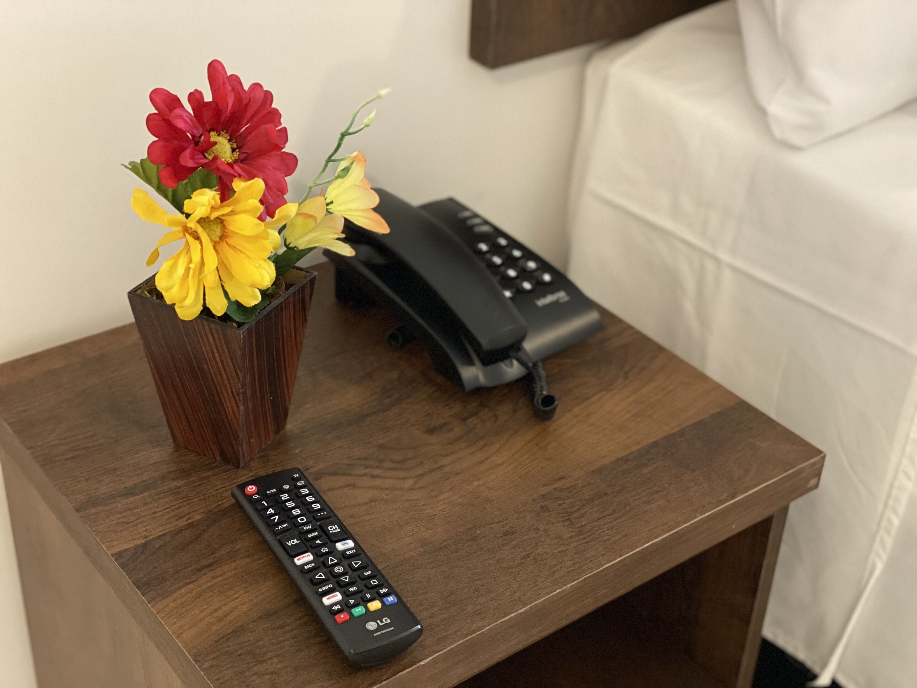 A wooden nightstand with a vase of flowers and a remote control.