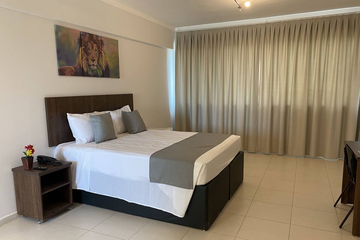 A modern hotel room with a large bed, a wooden headboard, a bedside table with a phone and remote, a painting on the wall, and a view of the outside through a curtain.