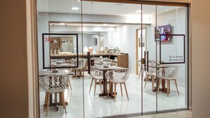 Interior de un restaurante con sillas y mesas blancas, y un área de cocina visible a través del vidrio.
