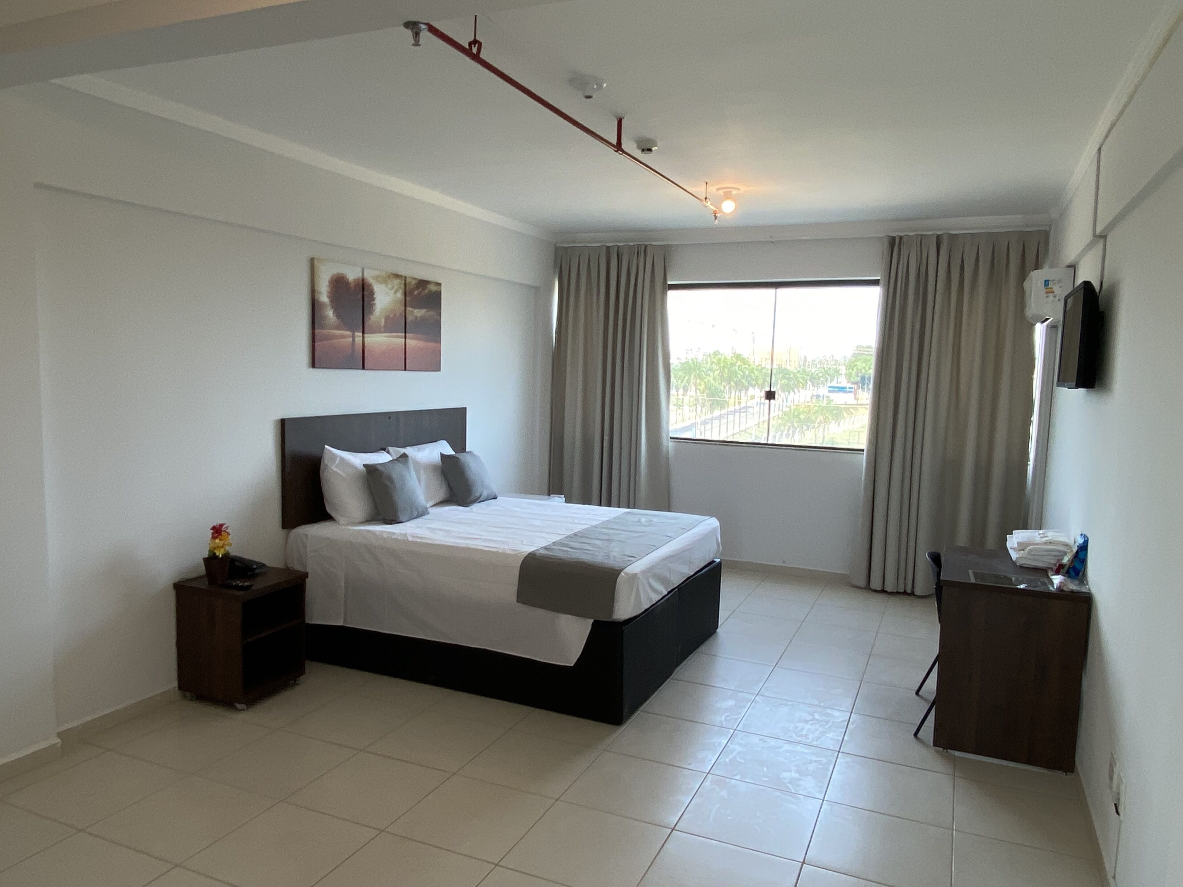 A bedroom with a bed, a nightstand, a dresser, a window with curtains, and a ceiling light fixture.