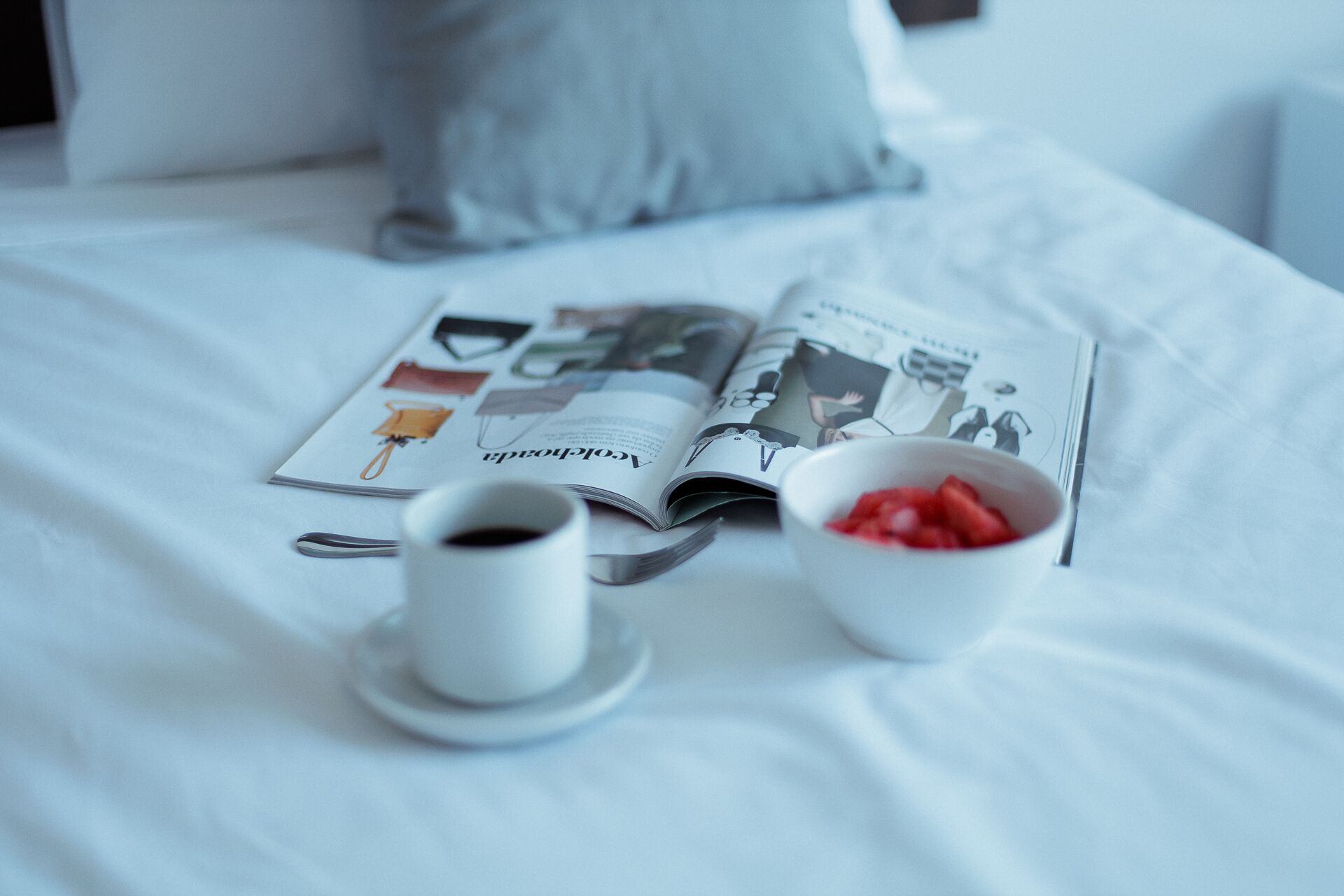 A bed with a magazine, a cup of coffee, and a bowl of strawberries.