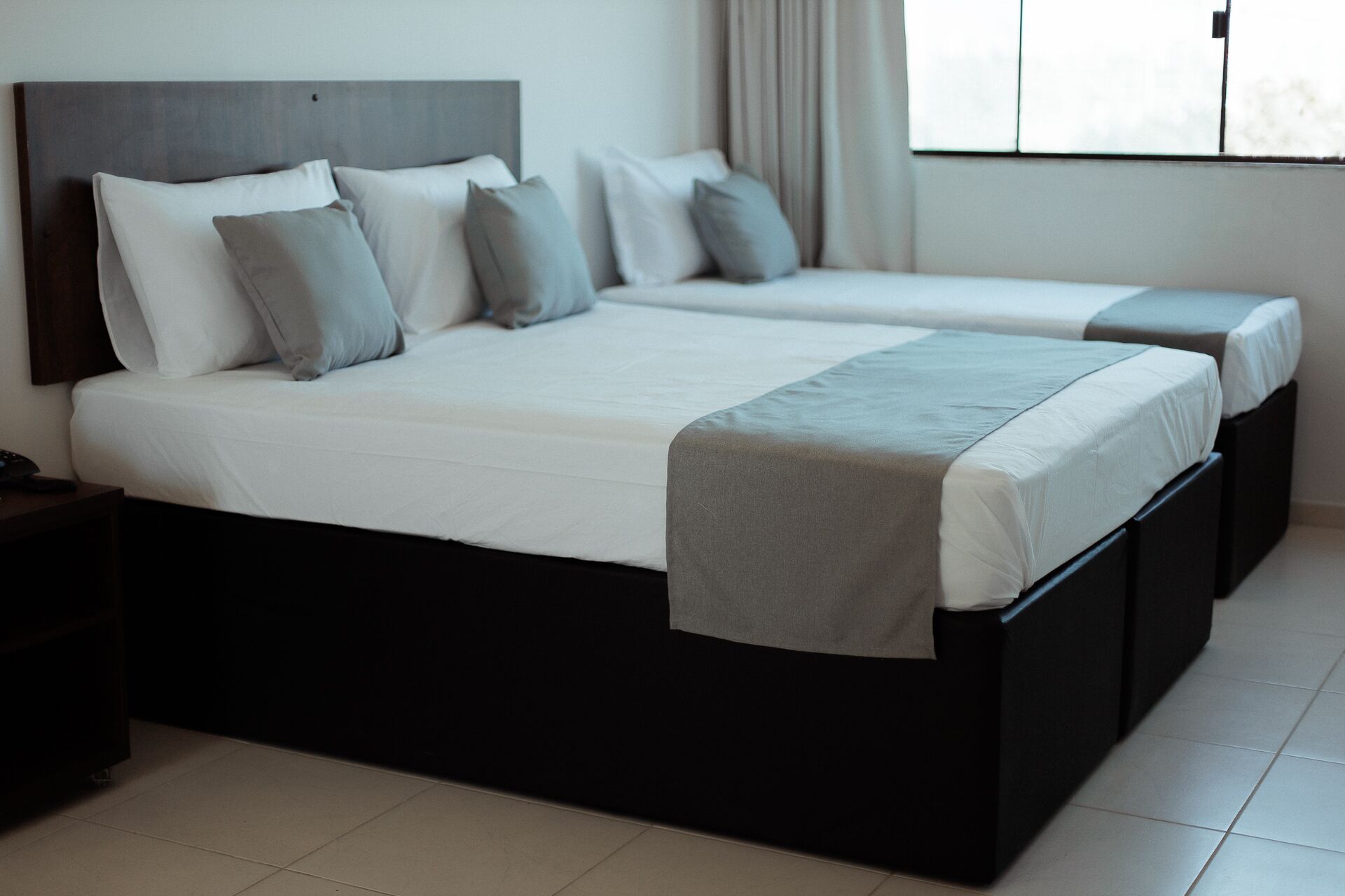 A double bed with a grey headboard and white bedding, a bedside table with a phone, and a window with curtains.