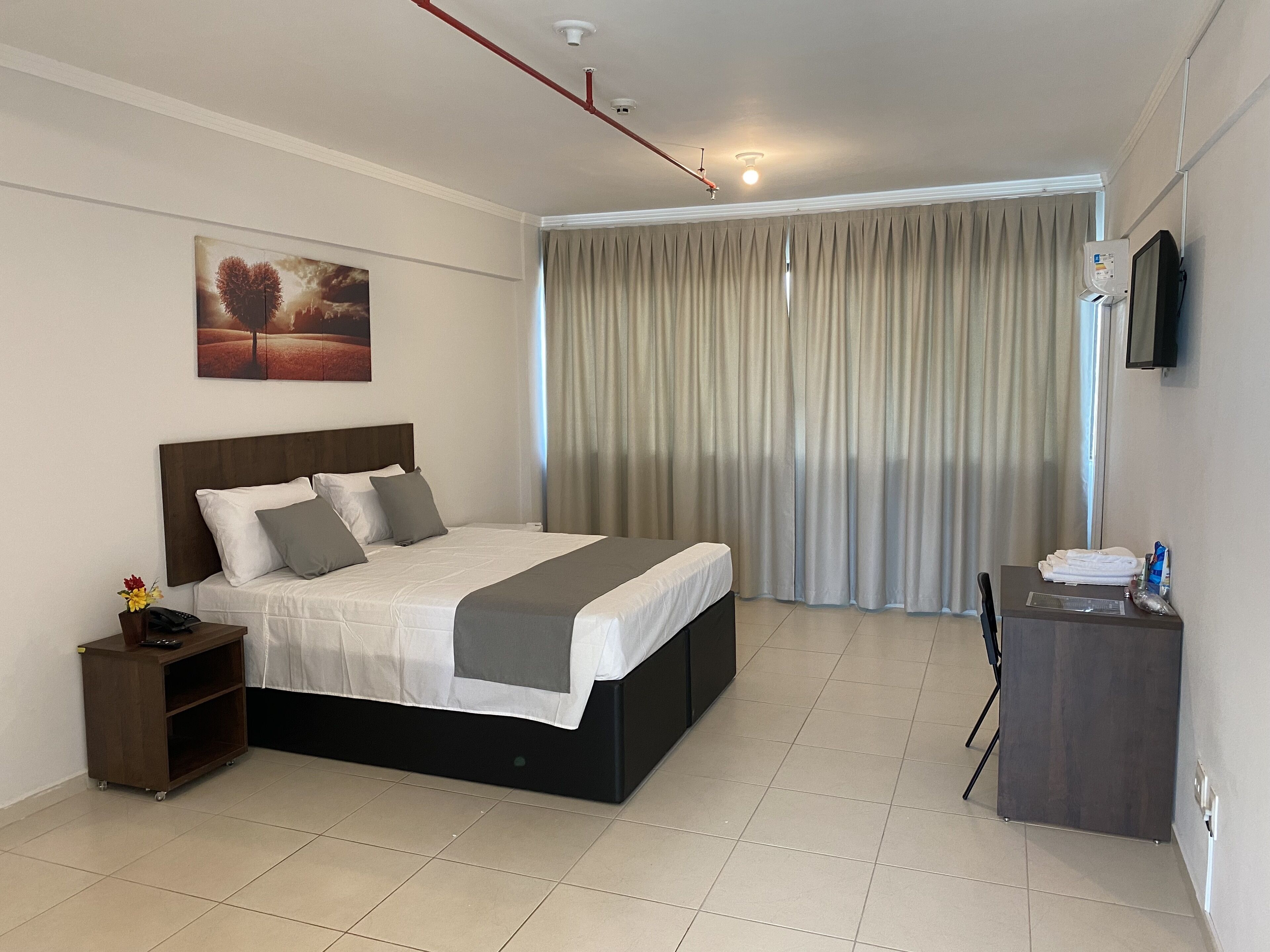 Una habitación de hotel con cama, escritorio, silla, televisión y ventana con cortinas.