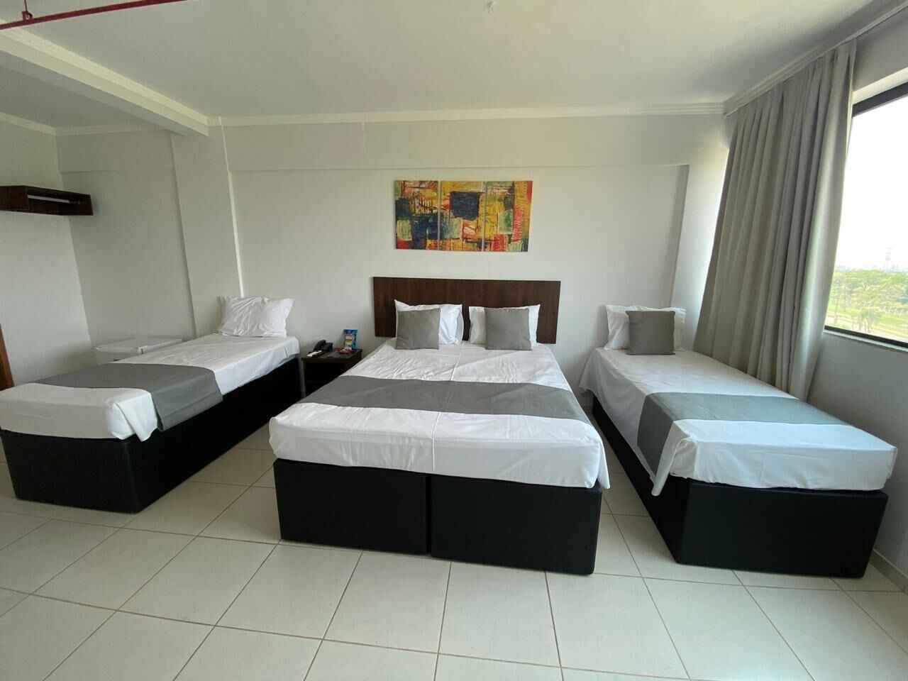 A hotel room with three beds arranged in a U-shape, a large window with curtains, and a colorful abstract painting on the wall.