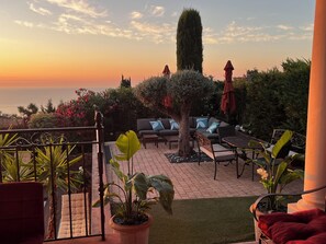 Une terrasse avec des meubles d’extérieur, une plante en pot et une vue sur la mer au coucher du soleil.