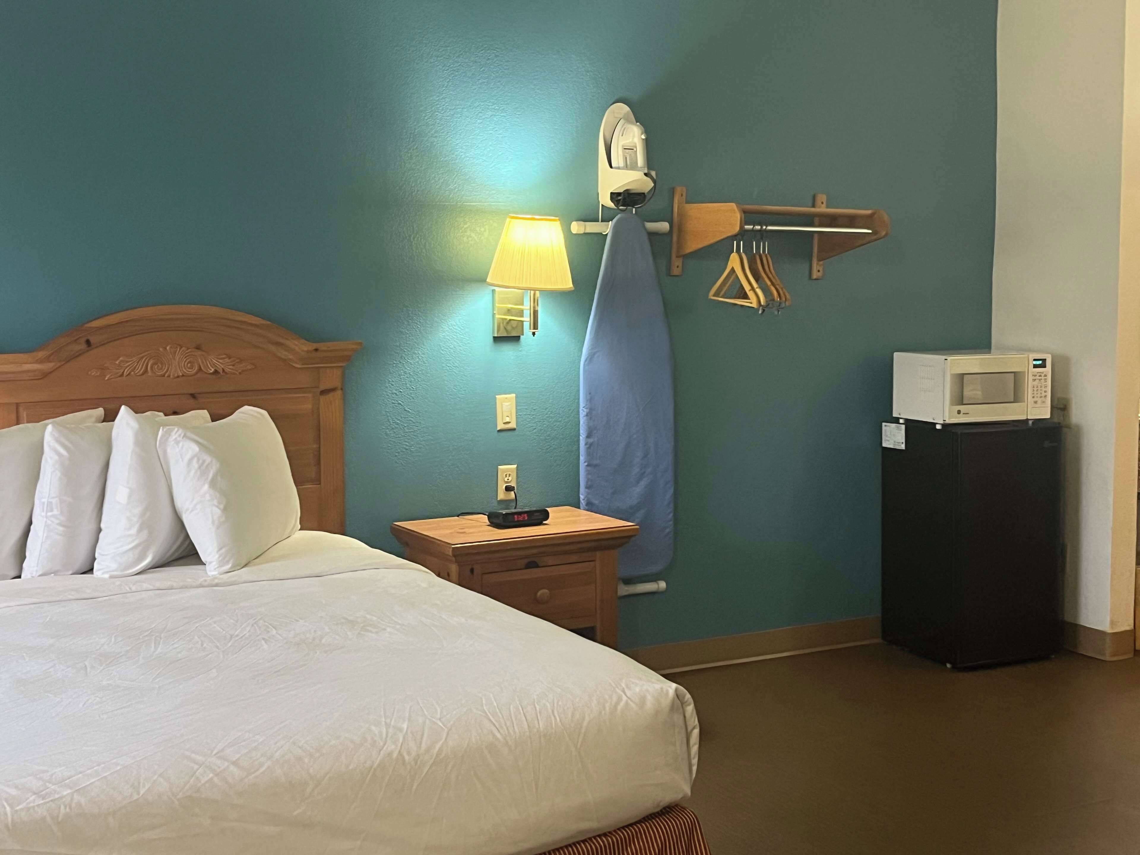 A hotel room with a bed, a nightstand, a lamp, an ironing board, a microwave, and a refrigerator.