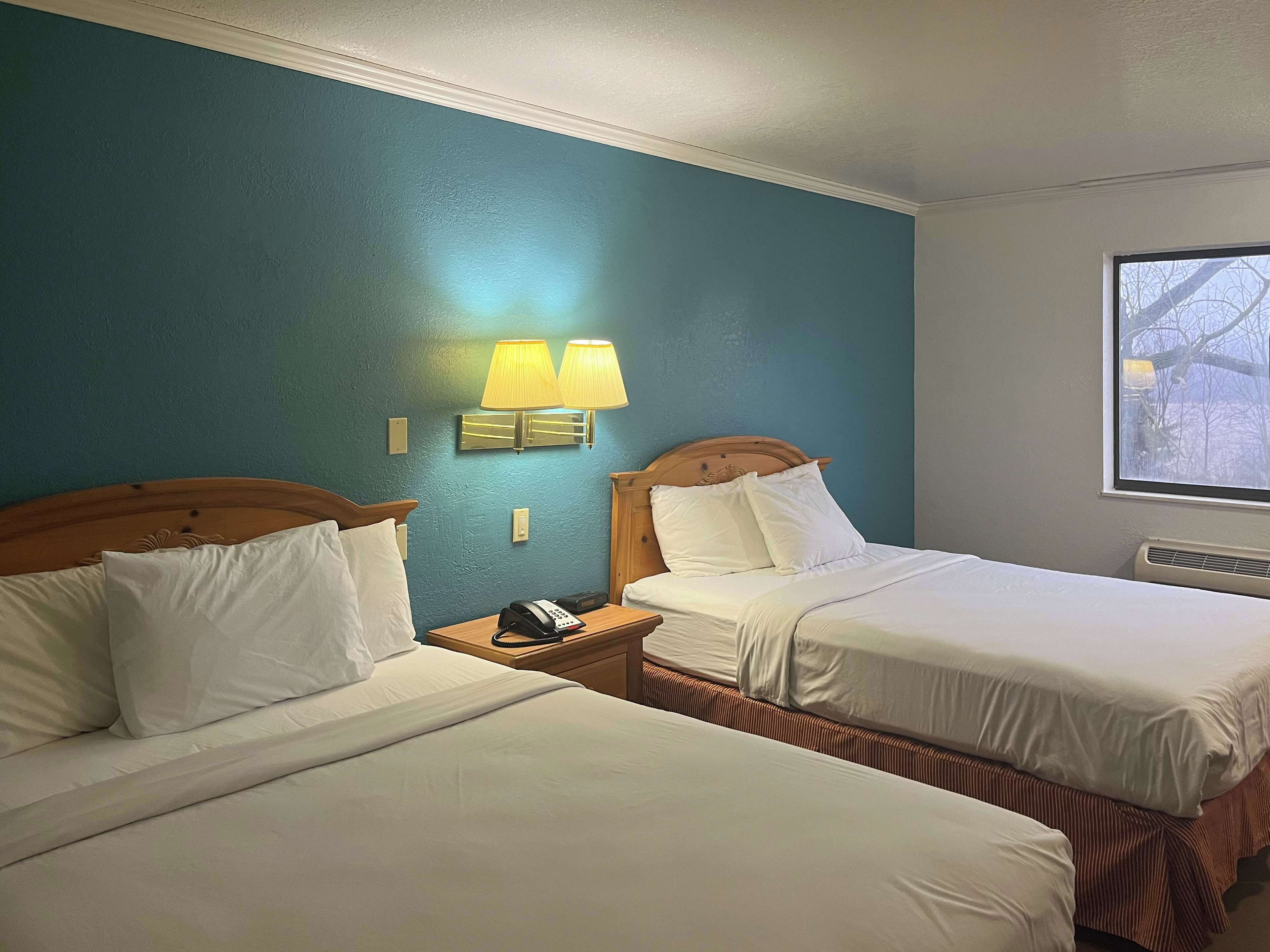 A hotel room with two beds, a nightstand with a phone, and a window with a view of trees.