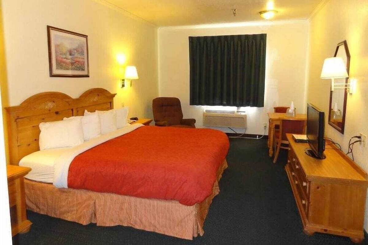 A hotel room with a large bed, a desk with a computer, a chair, and a window with curtains.