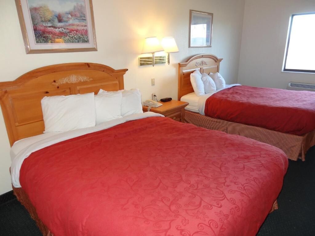 Quarto de hotel com duas camas, cada uma com colcha vermelha e almofadas brancas. Há uma pintura na parede, um abajur na mesa de cabeceira e uma janela que oferece luz natural.