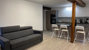 Un salon moderne avec un canapé gris et des chaises pliantes blanches, un plafond avec poutres apparentes en bois, et un coin cuisine doté de meubles gris et d’un four à micro-ondes.