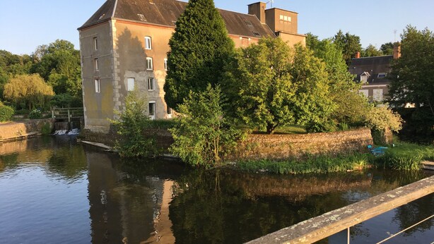 Un canal surmonté d’un pont en pierre, un grand bâtiment doté de nombreuses fenêtres et une végétation luxuriante.