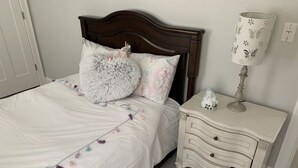 Un lit bien fait, avec une couette blanche et des coussins décoratifs, une tête de lit en bois, une table de chevet blanche avec une lampe, et un tapis à motifs.