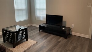 Een woonkamer met een tv op een standaard, twee ramen met zonwering, een glazen salontafel en een donkere houten vloer.