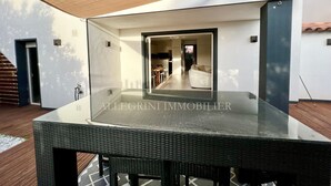 A modern outdoor seating area with a glass table and wicker chairs, a wooden deck, and a view into the house. - Beautiful villa 10 min walk from Ile Rousse (monticello)