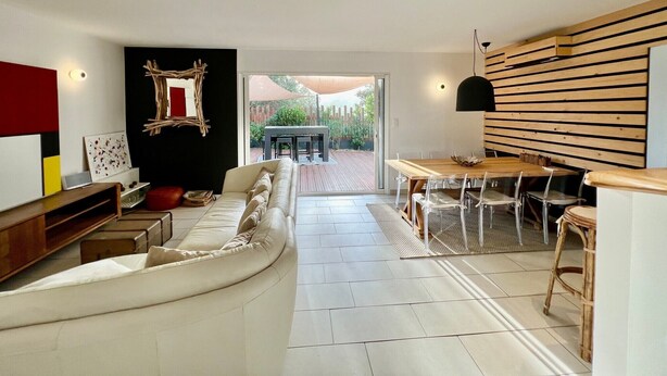 A modern living room with a sofa, a dining table with chairs, a wooden wall panel, and a view of the outdoor patio. - Beautiful villa 10 min walk from Ile Rousse (monticello)