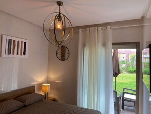 A bedroom with a bed, a bedside lamp, a wall clock, and a view of the garden through a sliding door. - Luxury Apartment - Prestigia Marrakech Golf City (Marrakech)