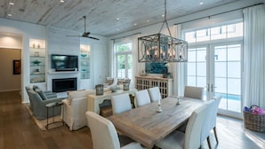 A spacious dining area with a wooden table and white chairs, a television mounted on the wall, and a large window with curtains. - 92 East Water Street | 4 Bedroom with Pool & Hot Tub! (Inlet Beach)