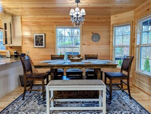 A wooden cabin interior with a dining table set for six, surrounded by chairs, a chandelier, and large windows offering a view of the outdoors.