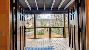 Une terrasse en bois avec un tapis à motifs bleus, de grandes fenêtres offrant une vue sur un espace paysager agrémenté d’arbres et sur une chaîne de montagnes lointaine.