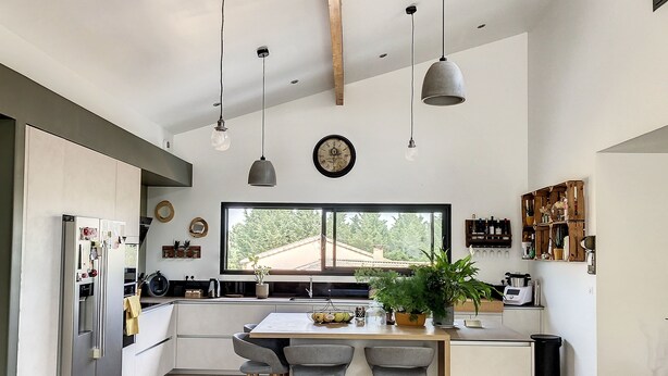 A modern kitchen with a large island, bar stools, hanging pendant lights, and a view of the outdoors through a large window.