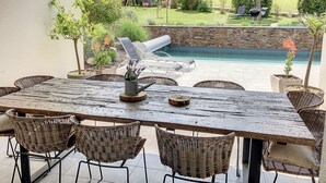 A patio with a dining table, chairs, a grill, and a view of a swimming pool and garden.