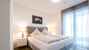 A neatly made bed with white linens, two bedside tables with lamps, a large window with sheer curtains, and a feather artwork on the wall. - Beary - modern ground floor flat in best location (Garmisch-Partenkirchen)