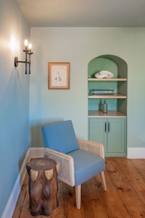 A room with a blue chair, a wooden stool, a green cabinet, and a wall-mounted light fixture.