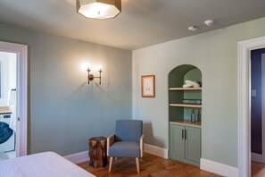 A bedroom with a bed, a chair, a wooden stool, a wall-mounted light fixture, a framed picture, and a green cabinet with shelves.