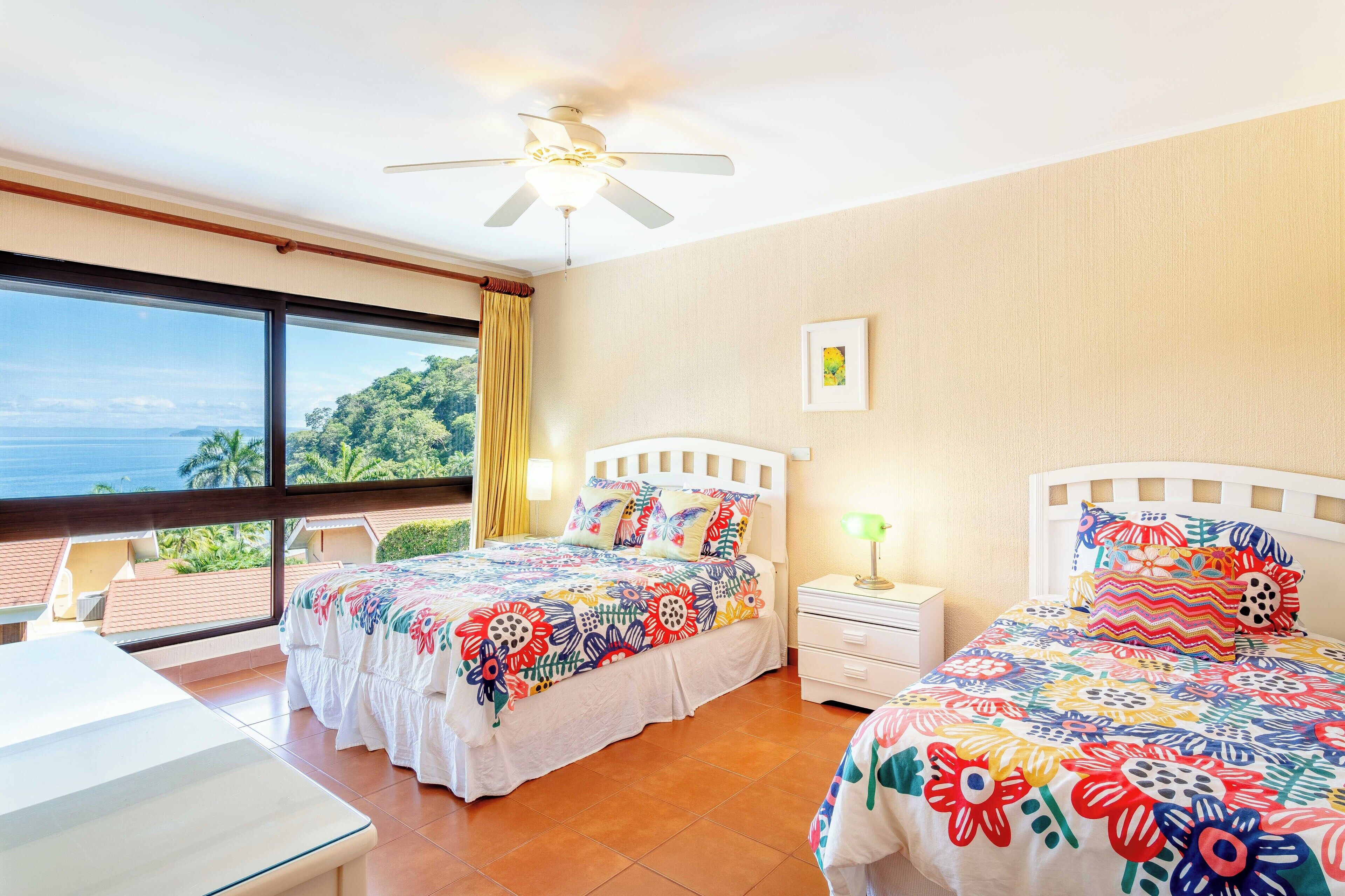 A hotel room with two beds, a ceiling fan, a large window with a view of greenery, and a small bedside table.