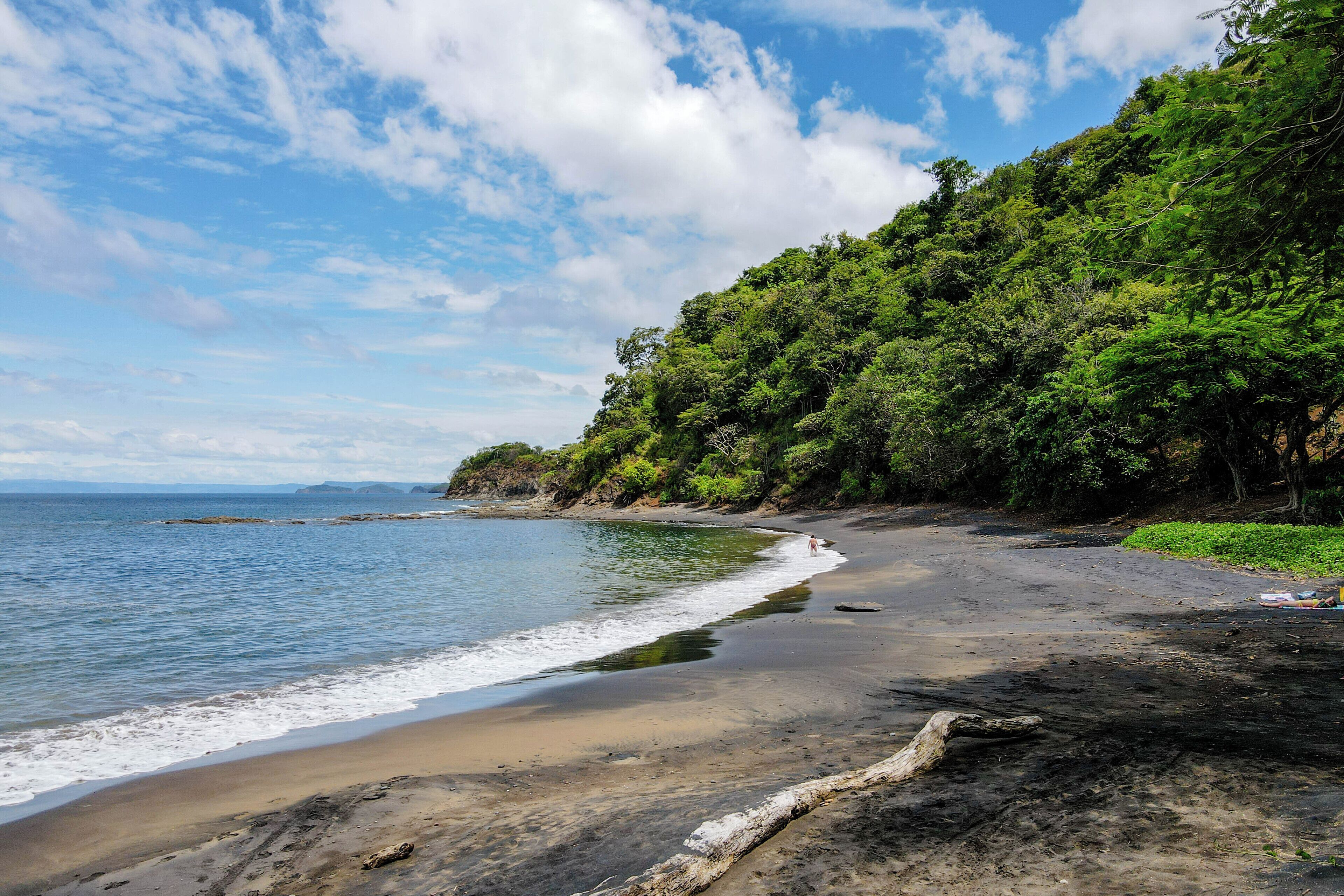 A beach with a rocky shoreline, a person walking along the shore, and dense green foliage on the hillside.