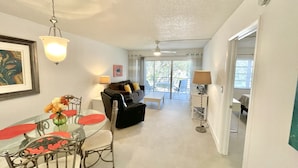 A spacious living area with a dining table, a sofa, and a lamp. There is a ceiling fan and a window with curtains.