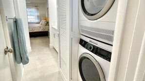 A laundry room with two stacked front-loading washing machines, a bed with a window, and a door with a towel hanging on it.