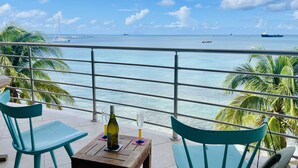 A balcony with turquoise chairs and a small table, overlooking a calm sea with boats and a ship in the distance.