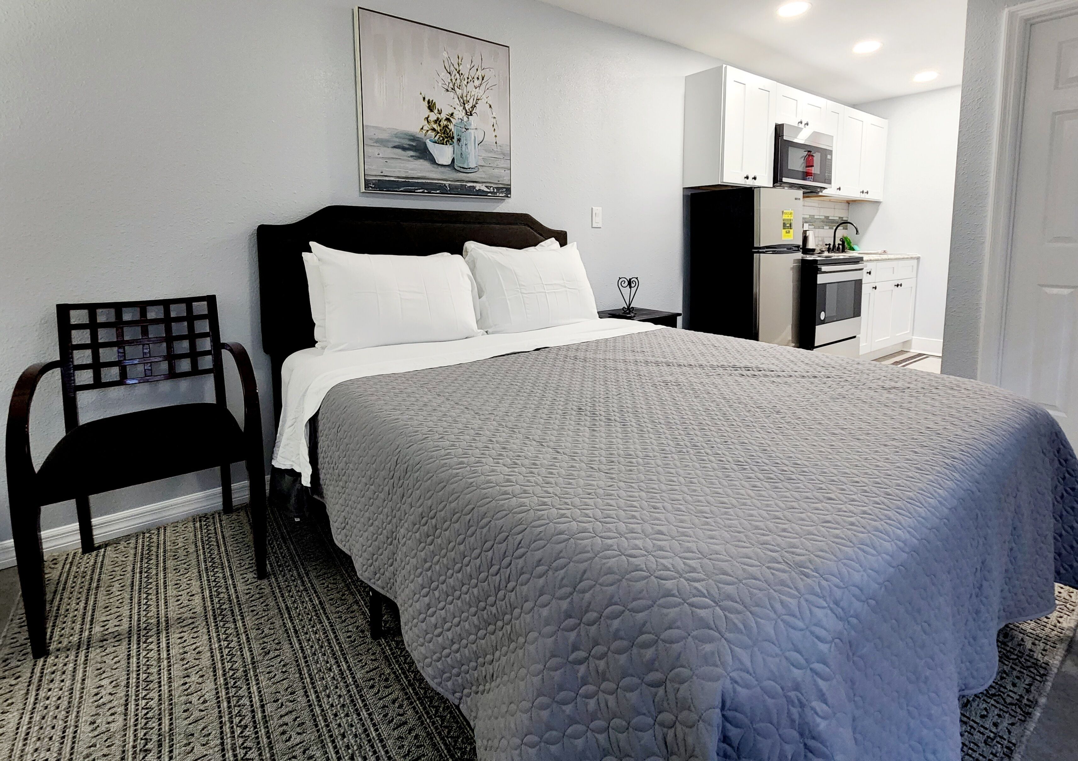 Un dormitorio ordenado con una cama grande, una silla, un cuadro y una kitchenette al fondo.