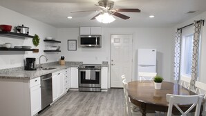 A modern kitchen with white cabinets, a stainless steel dishwasher, a microwave, and a built-in oven. There is a dining table with chairs and a ceiling fan above. - Modern 3-bedroom Guest Suite by Silicon Slopes (Eagle Mountain)