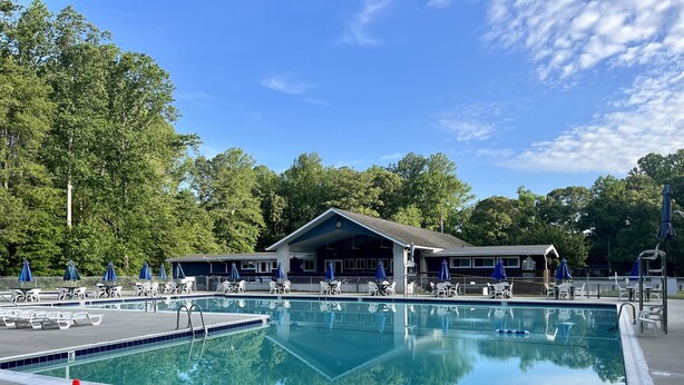 A rectangular swimming pool with a lifeguard stand, surrounded by lounge chairs and umbrellas, with a building in the background. - Beach House in waterfront gated community w/ salt water pool (Lewes)