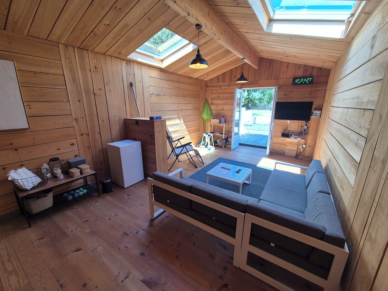 Interior de una cabaña de madera con sofá, una mesita, refrigerador y tragaluz.