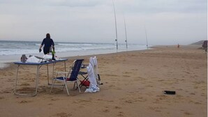 Ein Strand mit Tisch und Stühlen, eine Person geht spazieren, und Angelruten liegen im Sand.