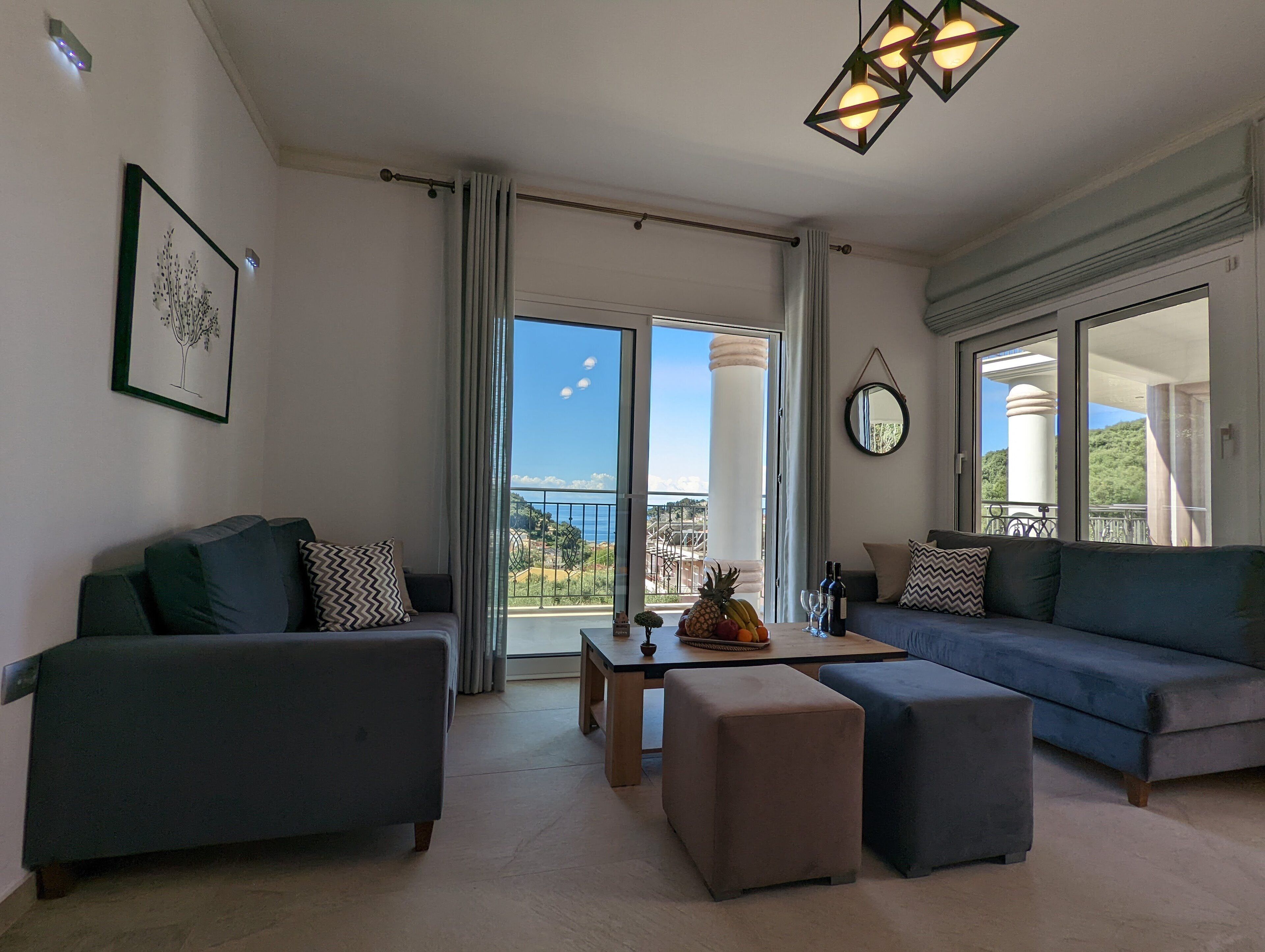 Sala de estar moderna com sofá, banqueta e uma mesa com frutas e vinho. Uma grande janela oferece vista para o exterior.