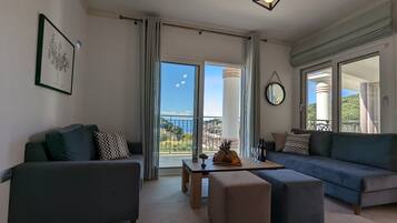 Sala de estar moderna com sofá, banqueta e uma mesa com frutas e vinho. Uma grande janela oferece vista para o exterior.