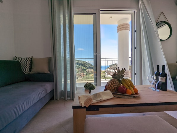 A balcony with a view of the sea, a table with fruits and wine bottles, and a sofa with cushions. - Natura Luxury Suites Parga (Parga)