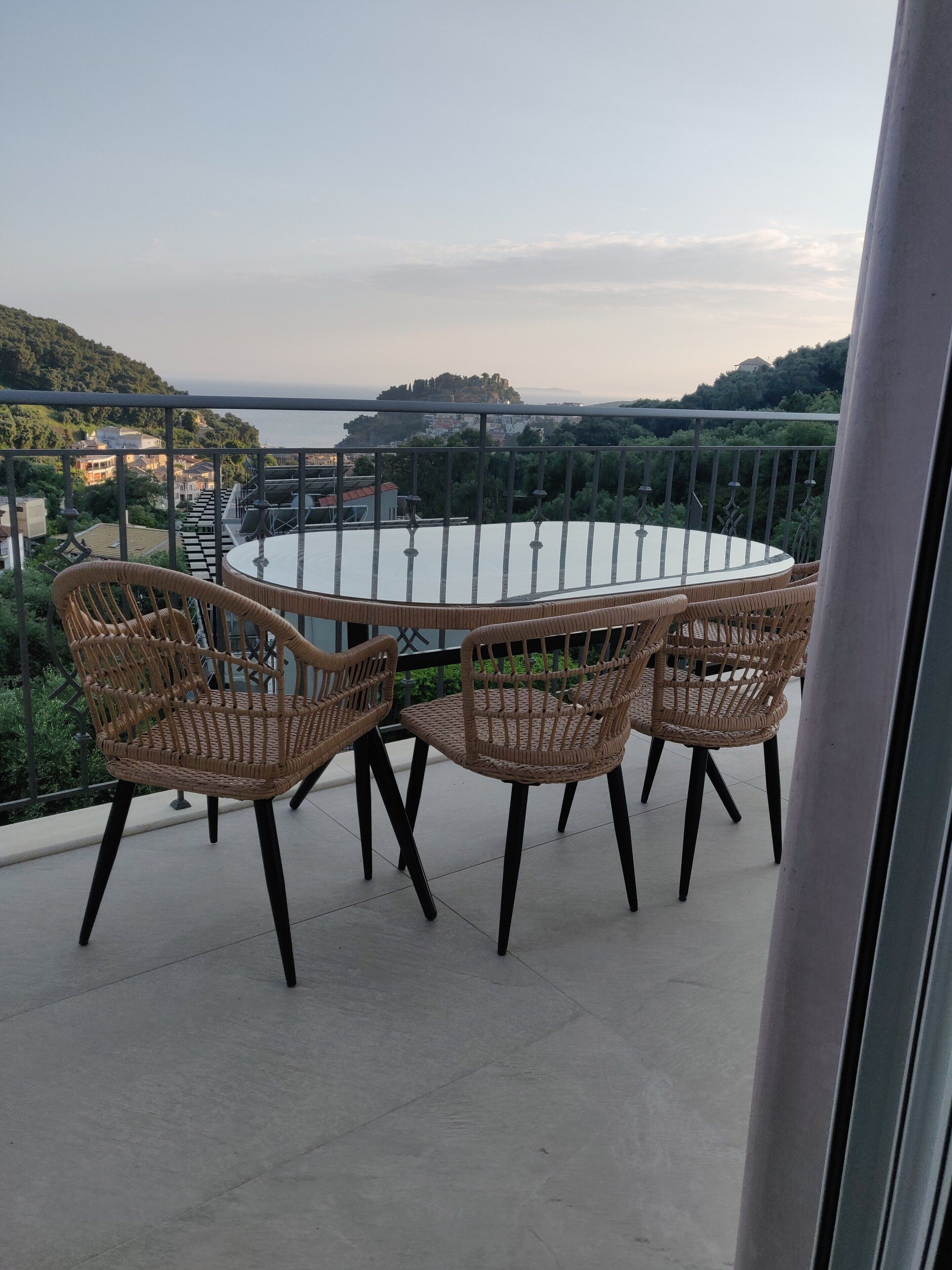 Un balcón con sillas de mimbre y una mesa de vidrio, con vistas a un paisaje costero con edificios y vegetación.