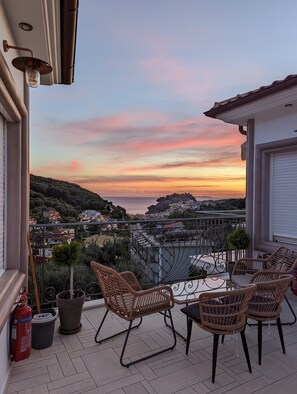 A balcony with wicker chairs, a table, and a view of a coastal town at sunset. - Natura Luxury Suites Parga (Parga)
