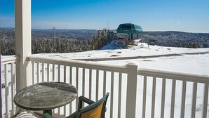 Ein mit Schnee bedeckter Balkon mit Tisch und Stuhl, mit Blick auf eine Seilbahn und bewaldete Berge.