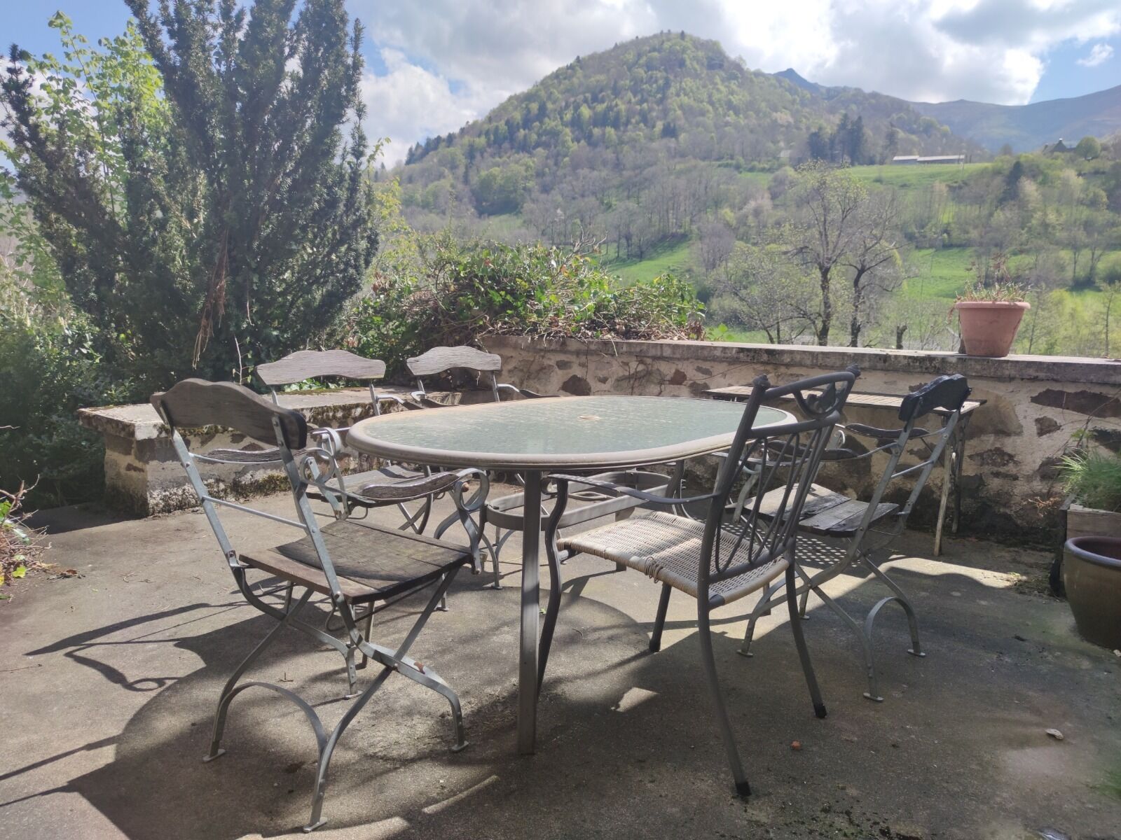 An outdoor patio with a round table and chairs, surrounded by stone walls and potted plants, overlooking a mountainous landscape.