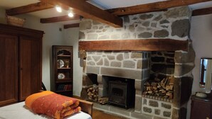 A bedroom with a stone fireplace, wooden beams, a bed with an orange blanket, a wooden cabinet, and a bookshelf.