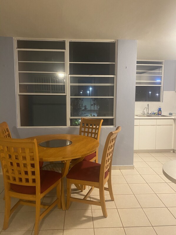 A dining area with a wooden table and chairs, large windows with blinds, and a kitchen area with a sink and cabinets. - cozy condo minutes from the beach - in the heart of Fajardo ❤️. (Farajdo)