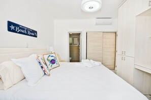 Quarto de hotel com cama, almofadas, uma placa com a inscrição 'Bem-vindo à nossa Beach House' e um guarda-roupa de madeira.